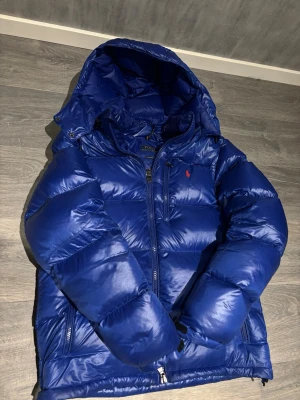 Blå dunjacka från Polo Ralph Lauren - Snygg blå dunjacka från Polo Ralph Lauren med huva och dragkedja framtill. Jackan har det klassiska röda Polo-logot på bröstet och två sidofickor med dragkedja. Perfekt för kalla dagar och riktigt skön att bära.