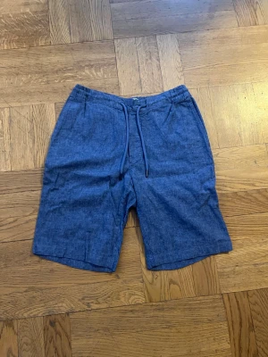 Blå linneshorts  - Snygga blå shorts i linnemix med snören i midjan och två sidofickor. Baktill finns två fickor med knappstängning. Perfekta för sommaren och riktigt sköna tack vare det lätta materialet.