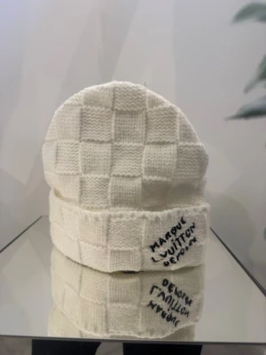 Vit Louis Vuitton stickad mössa - Exklusiv vit stickad mössa från Louis Vuitton med rutmönster och vikt kant. Tillverkad i en lyxig mix av 49% kashmir, 40% ull och 11% siden. Märkeslapp på insidan och broderad text på kanten för en unik touch.