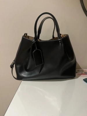 Zara väska - Stilren svart handväska i skinn med både handtag och justerbar axelrem. Väskan har vita kontrastsömmar och en dekorativ tag. Perfekt för dig som vill ha en klassisk och rymlig väska med snygga detaljer. Köpt på Zara i somras säljer pga att den aldrig kommer till användning. Använd 5 gånger.