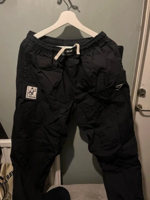 Svarta mjukisbyxor med patch och snörning  - Svarta mjukisbyxor/pumperpants från Gymshark  med vit snörning i midjan och patch med motiv på vänster ben. Byxorna har avslappnad passform och är perfekta för en chill stil. Materialet känns mjukt och bekvämt, och de har praktiska fickor framtill.