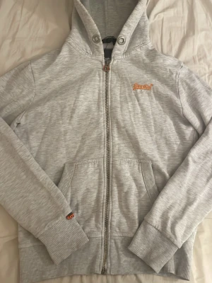 Ljusgrå hoodie från Superdry - Ljusgrå hoodie med dragkedja från Superdry. Har huva och snören tillkommer (sista bilden). Mycket bra skick bara lite nopprig