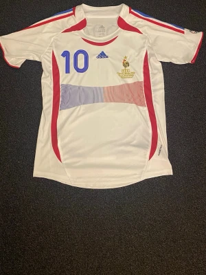 Frankrike Zidane fotbollströja Adidas - Säljer en vit Frankrike fotbollströja från Adidas med blå och röda detaljer, nummer 10 och Zidane på ryggen. Tröjan har korta ärmar, rund hals och tryck på både fram- och baksida. Perfekt för dig som älskar fotboll och klassiska landslagströjor. Den är väldigt stor så den är typ M