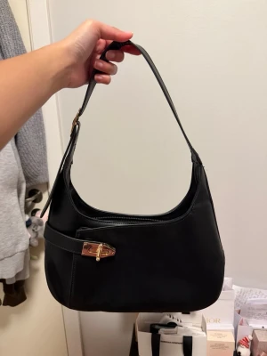 Svart axelväska med gulddetalj - Stilren svart axelväska i en klassisk baguetteform med guldfärgad spänndetalj framtill. Väskan har en enkel design med justerbart axelband och dragkedja upptill. Materialet ser ut att vara slätt konstläder som ger en modern look.