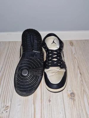 Nike Air Jordan 1 Low svart/vit - Snygga Nike Air Jordan 1 Low sneakers i svart och vitt med klassisk rund tå och svart sula. Skorna har snörning och ikoniska Jordan-loggan på plösen och hälen. Tillverkade i skinn och syntetmaterial för en stilren look. Skolådan ingår även