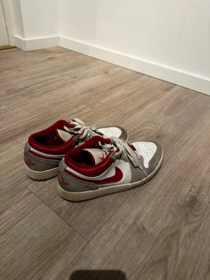 Nike Air Jordan 1 Low röd/vit/grå - Säljer ett par Nike Air Jordan 1 Low sneakers i vitt, grått och rött. Skorna har klassisk snörning, rund tå och den ikoniska swoosh-loggan på sidan. Ovandelen är i skinn och mocka med röda detaljer och perforerad tåbox för extra ventilation.