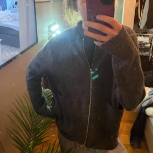 Mörkgrå stickad ziptröja - Mysig mörkgrå stickad tröja med dragkedja framtill och två fickor. Tröjan har en avslappnad passform och ribbstickade muddar vid ärmslut och nederkant. Perfekt för lager-på-lager och chill dagar.