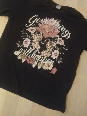 Svart t-shirt med tigrar och blommor - Svart t-shirt med tryck av två tigrar, blommor och texten 'Good things will happen' i vitt och rosa. T-shirten har rund hals och korta ärmar. Perfekt för dig som gillar coola och inspirerande motiv.