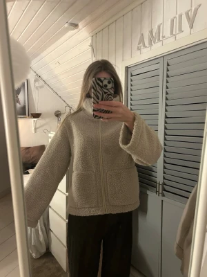 Beige teddyjacka med dragkedja - Mysig beige teddyjacka med hög krage och dragkedja framtill. Jackan har två stora fickor och är tillverkad i ett fluffigt teddy-material som ger en avslappnad och trendig look. Perfekt för kyliga dagar och enkel att matcha med olika outfits.