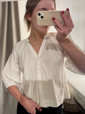 Vit blus/skjorta med 3/4-ärm och v-ringning - Stilren vit blus i luftig bomull med v-ringning och knäppning framtill. Blusen har 3/4-ärmar med lätt puff och avslappnad passform. Perfekt för en trendig och enkel look.