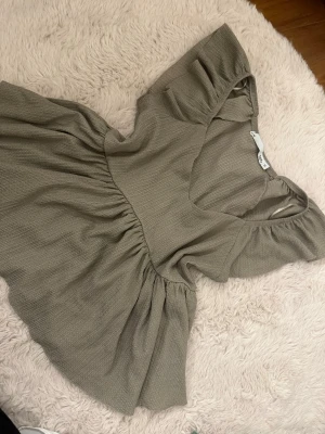 Brun blus - Superfin brun blus med v-ringning och korta ärmar. Blusen har en lös passform med volang nedtill och är tillverkad i ett mjukt bomullsliknande material. Perfekt för en avslappnad och trendig look.