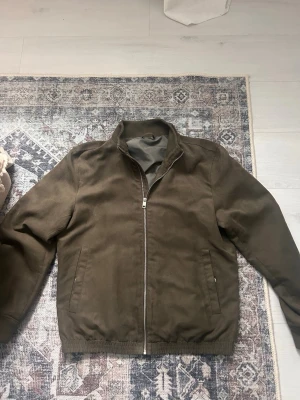 Olivgrön mocka jacka  - Snygg olivgrön jacka med rak passform och ståkrage. Jackan har dragkedja framtill, två sidofickor och elastisk nederkant. Materialet ser ut att vara bomull eller en bomullsblandning, perfekt för lager på lager under kallare dagar.