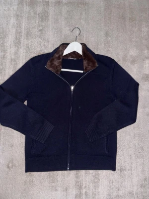 Merino kofta med pälskrage - Snygg svart ziptröja med mjuk brun fuskpäls i kragen. Tröjan har långa ärmar, ribbade muddar och dragkedja framtill. Perfekt för dig som vill ha en stilren men ändå unik look.