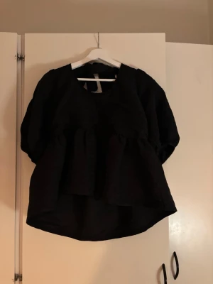 Svart volangblus med puffärm - Säljer en svart blus med volang och puffiga trekvartsärmar. Toppen har en v-ringad hals och knyt i nacken för extra detalj. Passformen är loose och tyget känns lätt och luftigt, perfekt för dig som gillar statement-ärmar.