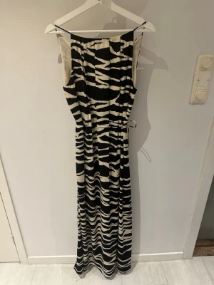 Svartvit långklänning med zebramönster - Cool långklänning med svartvitt zebramönster och ärmlös design. Klänningen har en rak passform och är tillverkad i ett mjukt, följsamt material som känns skönt mot huden. Perfekt för dig som vill sticka ut med en trendig look. Smalt skärp i midjan.