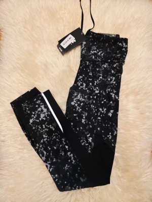 Svarta träningsleggings med mönster - Snygga svarta träningsleggings från Under Armour med grått och vitt pixelmönster. Hög midja och smal passform, samt vita reflexdetaljer längs benen. Perfekta för gymmet eller löprundan. Materialet är stretchigt och andas bra.