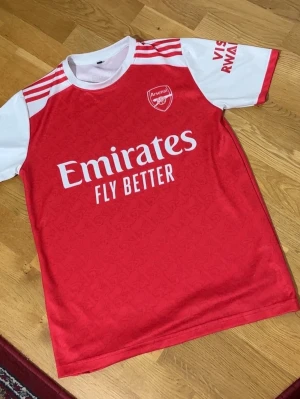 Arsenal matchtröja röd/vit - Snygg Arsenal matchtröja i rött och vitt med klubbmärke på bröstet och 'Emirates Fly Better' tryckt på framsidan. Tröjan har vita ärmar med röda detaljer och rund hals. Perfekt för dig som älskar fotboll och vill stötta ditt lag. Storlek: S-M