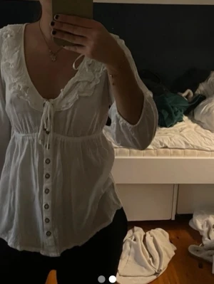 Vit blus med volang och knappar H&M - Söt vit blus från H&M med volangdetaljer runt den djupa ringningen och knytband framtill. Blusen har knappar längs hela framsidan och trekvartsärm. Tillverkad i ett lätt tyg som ger en luftig känsla. mo