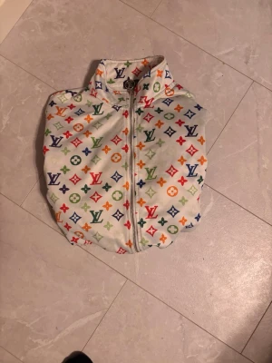Färgglatt Louis Vuitton set - Säljer ett vitt set med dragkedja och byxa från Louis Vuitton, täckt av deras ikoniska monogram i flera färger som blått, rött, grönt och orange. Setet har randiga muddar och är gjort i ett mjukt, glansigt material. Perfekt för dig som vill sticka ut med en unik look.