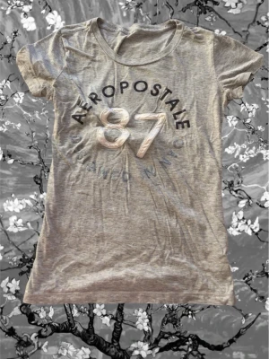 Grå Aeropostale t-shirt med tryck - Snygg grå t-shirt från Aeropostale med stort tryck framtill där det står '87' och 'AEROPOSTALE'. T-shirten har rund halsringning och korta ärmar. Tillverkad i mjuk bomull för en skön känsla och en slim passform som sitter snyggt.