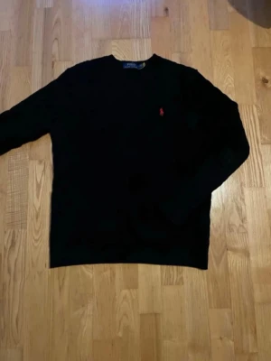 Svart stickad tröja Polo Ralph Lauren - Svart stickad tröja från Polo Ralph Lauren med rund halsringning, mönster och klassisk röd broderad logga på bröstet. Tröjan har lång ärm och ribbade muddar vid ärmslut och nederkant. Perfekt för dig som gillar stilrena och tidlösa plagg.