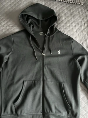 Svart hoodie från Polo Ralph Lauren - Svart hoodie med dragkedja från Polo Ralph Lauren. Klassisk design med vit broderad logga på bröstet, huva med dragsko och två stora fickor framtill. Tillverkad i mjukt material som är skönt att bära. Perfekt för en avslappnad och stilren look.