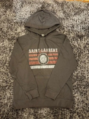 Grå hoodie från Saint Laurent. Storlek M - Grå  hoodie från Saint Laurent med tryck framtill i vitt och rött. Klassisk huva med dragsko och känguruficka. Mjukt material och ribbade muddar vid ärmslut och nederkant. Perfekt för en avslappnad och trendig look.