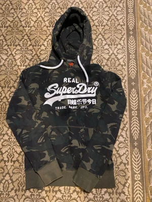 Camouflage hoodie från Superdry M - Snygg hoodie från Superdry i mörkgrönt camouflage-mönster med stor vit logga på bröstet. Hoodien har justerbar snörning i huvan och en klassisk känguruficka framtill. Tillverkad i mjuk bomullsmix, perfekt för en avslappnad streetwear-look. Hoodien är lite trasig vid halsen.