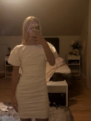 Vit kort puffärmad klänning - Säljer en vit kort klänning med puffärmar och rynkade detaljer både fram och bak. Klänningen har en öppen rygg och en figurnära passform som framhäver siluetten. Perfekt för dig som gillar trendiga och stilrena plagg.