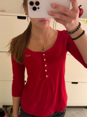 Röd henley topp 3/4 trekvarts ärm Hollister med knappar - Säljer en röd topp från Hollister med trekvartsärm och v-ringning. Toppen har vita knappar framtill och den klassiska Hollister-loggan broderad på bröstet. Perfekt till jeans eller kjol för en enkel men snygg look.