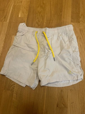 Beige badbyxor från Gant med gula detaljer - Snygga beige badbyxor från Gant med gula dragsnören och broderad logga på benet. Klassisk modell med elastisk midja och sidofickor. Tillverkade i snabbtorkande material och har meshfoder på insidan för extra komfort.