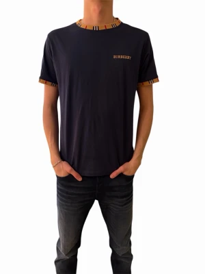 Mörkblå Burberry t-shirt med ränder - Snygg mörkblå t-shirt från Burberry med klassiska randiga detaljer i beige, svart och rött vid halsringning och ärmslut. T-shirten har korta ärmar och en liten Burberry-logga på bröstet. Tillverkad i mjuk bomull för en skön känsla.