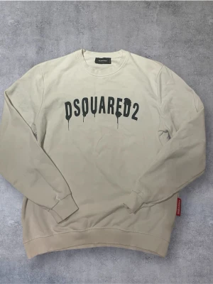 Cream white Sweatshirt från Dsquared2 - Cream white sweatshirt från Dsquared2 med svart tryck på bröstet och rund hals. Tröjan har långa ärmar, ribbade muddar och en röd etikett med logga i nederkant. Perfekt för en avslappnad och trendig stil. Blå fläck på ena axeln ( e bild)