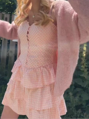 Ljusrosa prickig volangklänning - Supersöt ljusrosa kortklänning med vita prickar, smala axelband och knappar framtill. Klänningen har flera lager volanger nedtill och är gjord i ett lätt bomullstyg som känns perfekt för varma dagar.