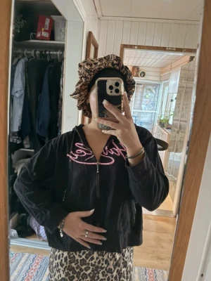 Svart Harley Davidsson hoodie med rosa tryck - Svart hoodie från Hurley med dragkedja och huva. På bröstet finns ett stort rosa Hurley-tryck i skrivstil. Hoodien har två fickor med dragkedja framtill och är tillverkad i mjukt material, perfekt för en chill och avslappnad stil. Sitter såhär på mig med storlek 40!