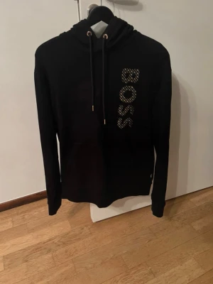 Svart hoodie från BOSS med tryck - Svart hoodie från BOSS med stort BOSS-tryck i vitt och guld på framsidan. Klassisk modell med huva och dragsnören med gulddetaljer. Perfekt för en stilren och trendig look. Helt ny endast använd en gång storlek S, nypris 2.7