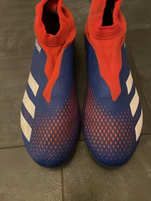 Adidas Predator blå/röda fotbollsskor - Säljer ett par Adidas Predator fotbollsskor i blått och rött med vita detaljer. Skorna har en strumpliknande passform utan snörning, mönstrad ovandel och fasta dobbar för spel på gräs. Ikoniska tre ränder på sidan och Predator-logga på hälen. Storlek 42-43