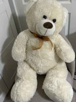 Stor vit nallebjörn med rosett - Mysig och fluffig stor nallebjörn i cremevit/beige plysch med en brun nos, svarta ögon och en ljusbrun rosett runt halsen. Perfekt som present eller dekoration i sovrummet. Nallen har en klassisk form och är riktigt mjuk att krama. Helt ny 