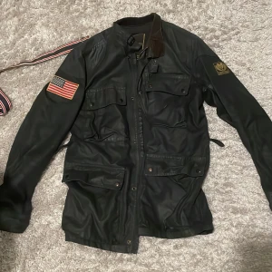 Svart jacka från Denim & Supply Ralph Lauren - Säljer en svart jacka från Denim & Supply Ralph Lauren med amerikansk flagga och broderad logga på ärmen. Jackan har flera fickor och knappar framtill, samt en bälte i midjan för justerbar passform.