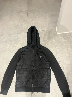 J.Lindeberg Cardigan🌟 - Svart cardigan jacka från J.Lindeberg med quiltad front och huva. Jackan har dragkedja framtill, två sidofickor med dragkedja och ribbade muddar. Ärmarna är i ett annat material än kroppen vilket ger en snygg kontrast. Perfekt för en stilren och sportig look. Vid snabb affär kan jag gå ner i pris!🌟👀