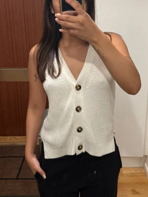 Vit stickad väst med knappar - Säljer en stilren vit stickad väst med breda axelband och djup V-ringning. Västen har fyra stora, mönstrade knappar framtill och är gjord i ett mjukt, ribbat material. Perfekt att styla över en t-shirt eller skjorta för en trendig look.