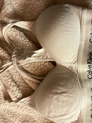 Beige sport-bh från Calvin Klein - Säljer en beige sport-bh från Calvin Klein med vadderade kupor och brottarrygg. Bh:n har bred resår med logotypen tryckt runt om och är tillverkad i mjukt bomullsmaterial med stretch. Perfekt för dig som gillar enkel och stilren design.