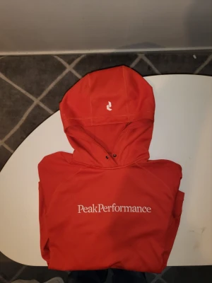 Orange hoodie från Peak Performance - Snygg orange hoodie från Peak Performance med vit logga framtill. Hoodien har huva med dragsko och långa ärmar. Tillverkad i ett mjukt och stretchigt material som passar perfekt för en aktiv livsstil. Enkel och stilren design med diskreta detaljer.
