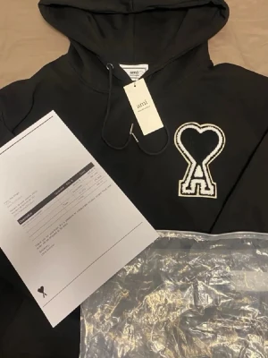 Svart hoodie från Ami Paris - Snygg svart hoodie från Ami Paris med stor vit broderad logga i form av ett hjärta och bokstaven A på bröstet. Klassisk huva med dragsko och mjukt material som känns skönt mot huden. Perfekt för en avslappnad och trendig look.