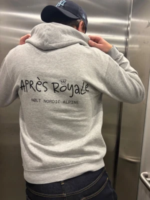 Grå hoodie Après Royale Nolt - Snygg grå hoodie från Après Royale med tryck på ryggen där det står 'Après Royale' och 'NOLT NORDIC ALPINE'. Klassisk passform med huva och mjukt material som känns skönt mot huden. Perfekt för dig som gillar stilrena och bekväma plagg.