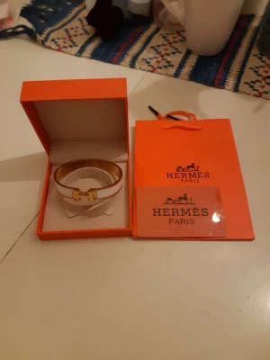 Vitt Hermès armband med roseguld  - Snyggt vitt armband från Hermès med guldfärgad H-detalj framtill. Kommer i original ask och med Hermès påse. Armbandet har en elegant och stilren design som passar perfekt till din accessoarsamling.