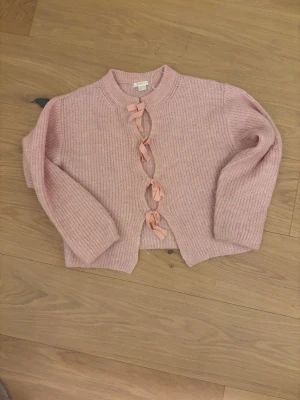 Rosa stickad kofta med rosetter - Supersöt rosa stickad kofta från Gina Tricot med tre dekorativa rosetter framtill. Koftan har rund halsringning, lång ärm och är gjord i ett mjukt, ribbstickat material. Perfekt för att addera en gullig touch till din outfit.