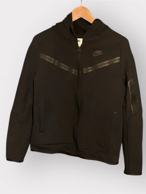 Svart Nike hoodie med dragkedja - Svart hoodie från Nike med hel dragkedja framtill och två snedställda fickor. Har en huva och diskreta svarta detaljer över bröstet samt en extra ficka på ena ärmen. Tillverkad i mjukt material, perfekt för en sportig och avslappnad stil.