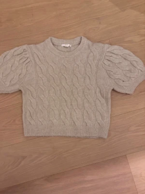 Beige kabelstickad tröja Gina Tricot - Beige kabelstickad tröja från Gina Tricot med korta puffärmar och ribbade muddar. Tröjan har en croppad passform och rund halsringning. Perfekt för lager-på-lager och en trendig look.