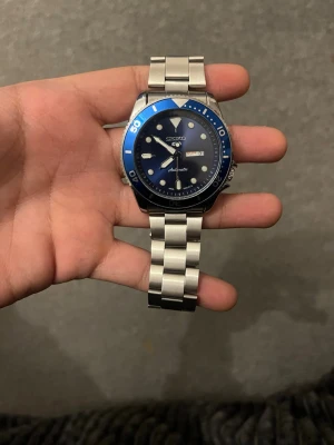 Seiko 5 Sports blå automatisk klocka - Snygg Seiko 5 Sports klocka med blå urtavla och blå vridbar bezel. Klockan har dag- och datumvisning samt robust länkarmband i rostfritt stål. Automatisk rörelse och tydliga indexmarkeringar gör den både stilren och funktionell.